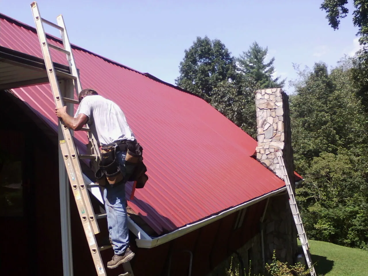 Roofer on a ladder beside a red metal roof and stone chimney — Soffit & Fascia Repair by Roofing Brothers in Saint Marys City